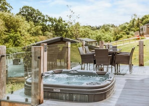 Un jacuzzi en una terraza de madera con sillas de mimbre y una mesa, rodeado de follaje.