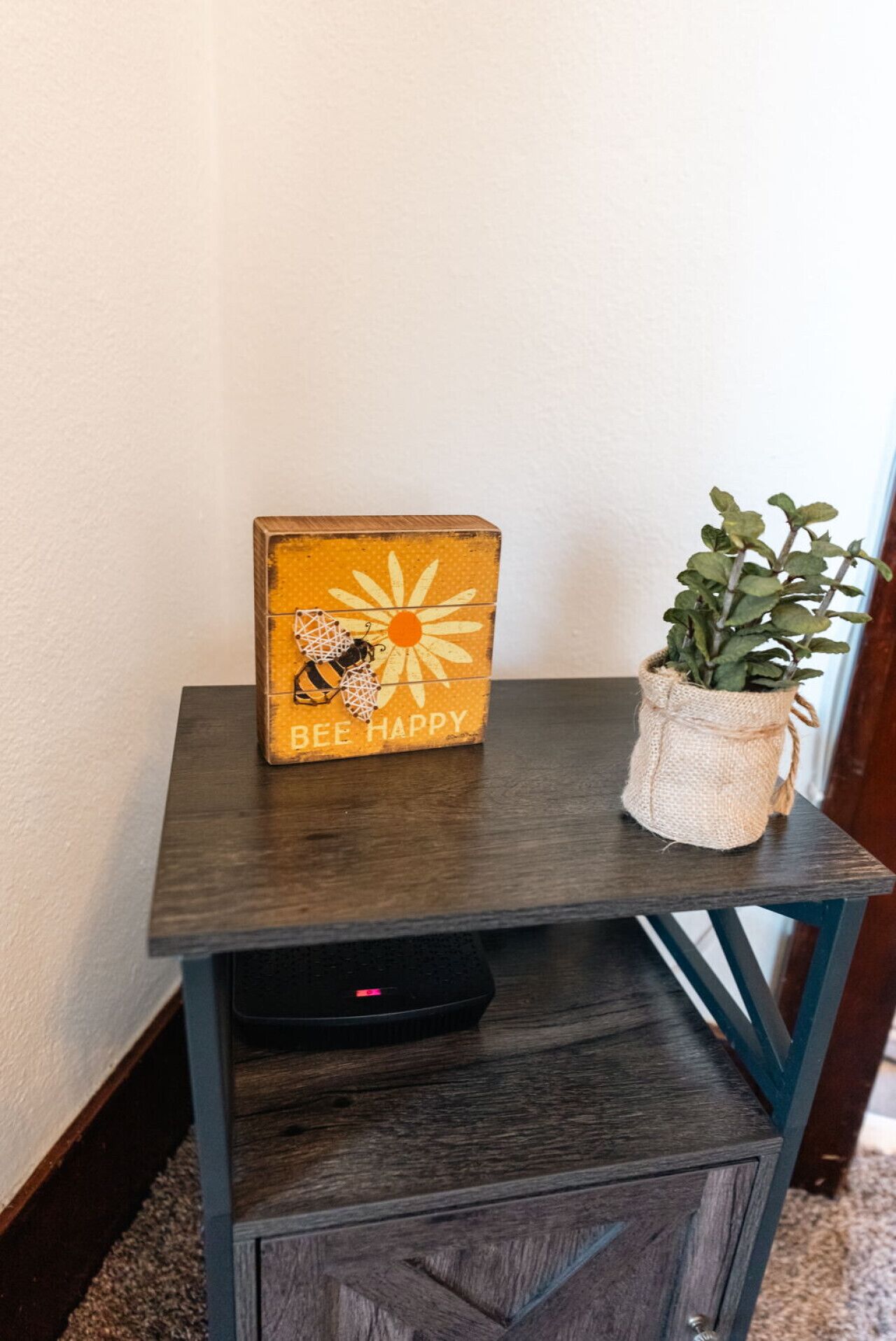 A small wooden table with a sign that reads "BEE HAPPY" and a potted plant on it.