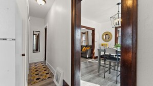 Un couloir mène à une salle à manger dont le sol est orné de motifs, où l’on trouve un luminaire moderne et un miroir accroché au mur.