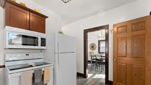 Une cuisine compacte équipée d’un four à micro-ondes, d’un four et d’un réfrigérateur. Un meuble en bois est placé au-dessus du four à micro-ondes, et une porte en bois se trouve sur le côté droit. Un tapis à motifs recouvre le sol.