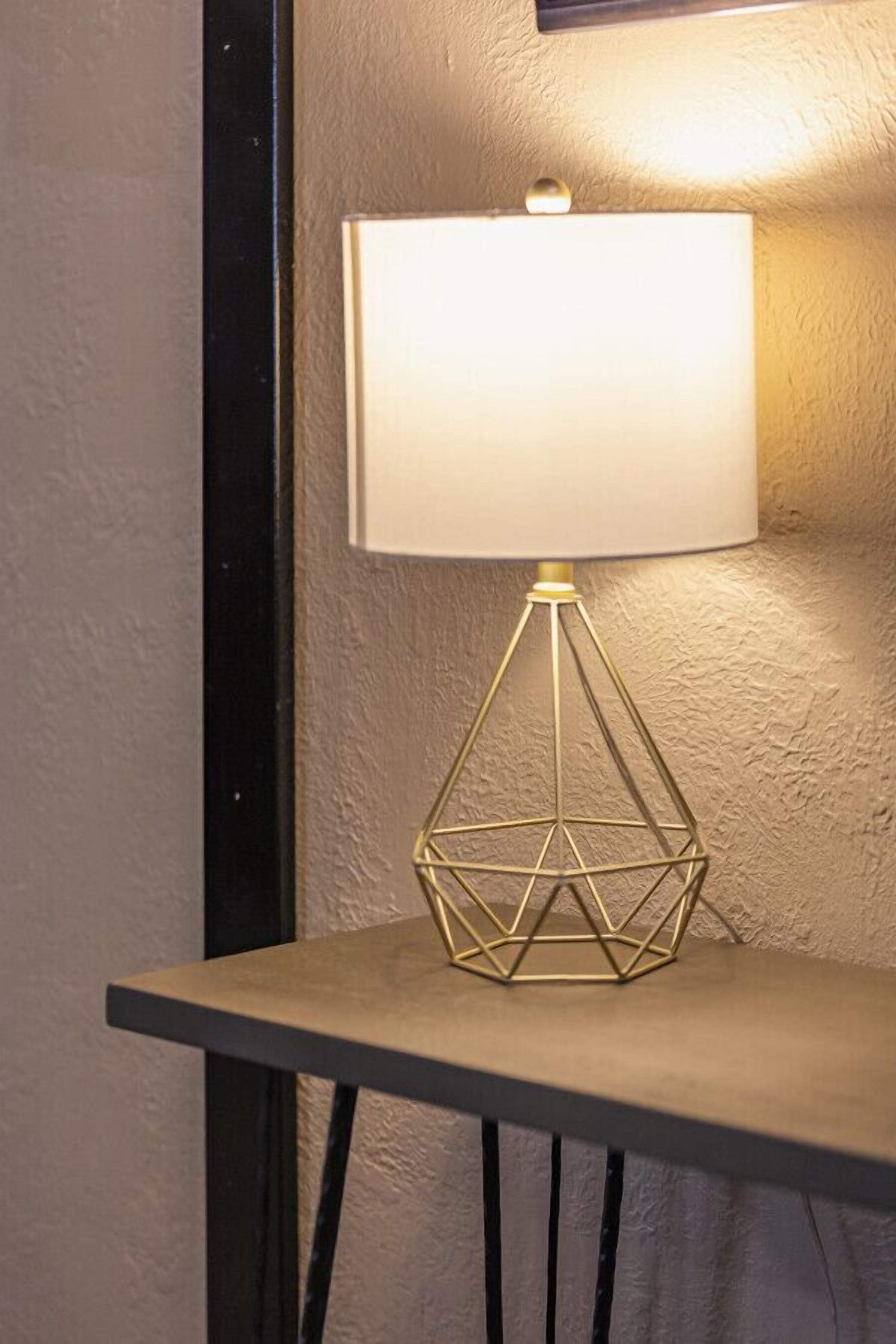 A table lamp with a geometric base and a white lampshade, placed on a table against a textured wall.