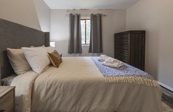 Una cama bien hecha con una colcha color beige y almohadones, una mesita de noche de madera, un armario de madera oscura y una ventana con cortinas.
