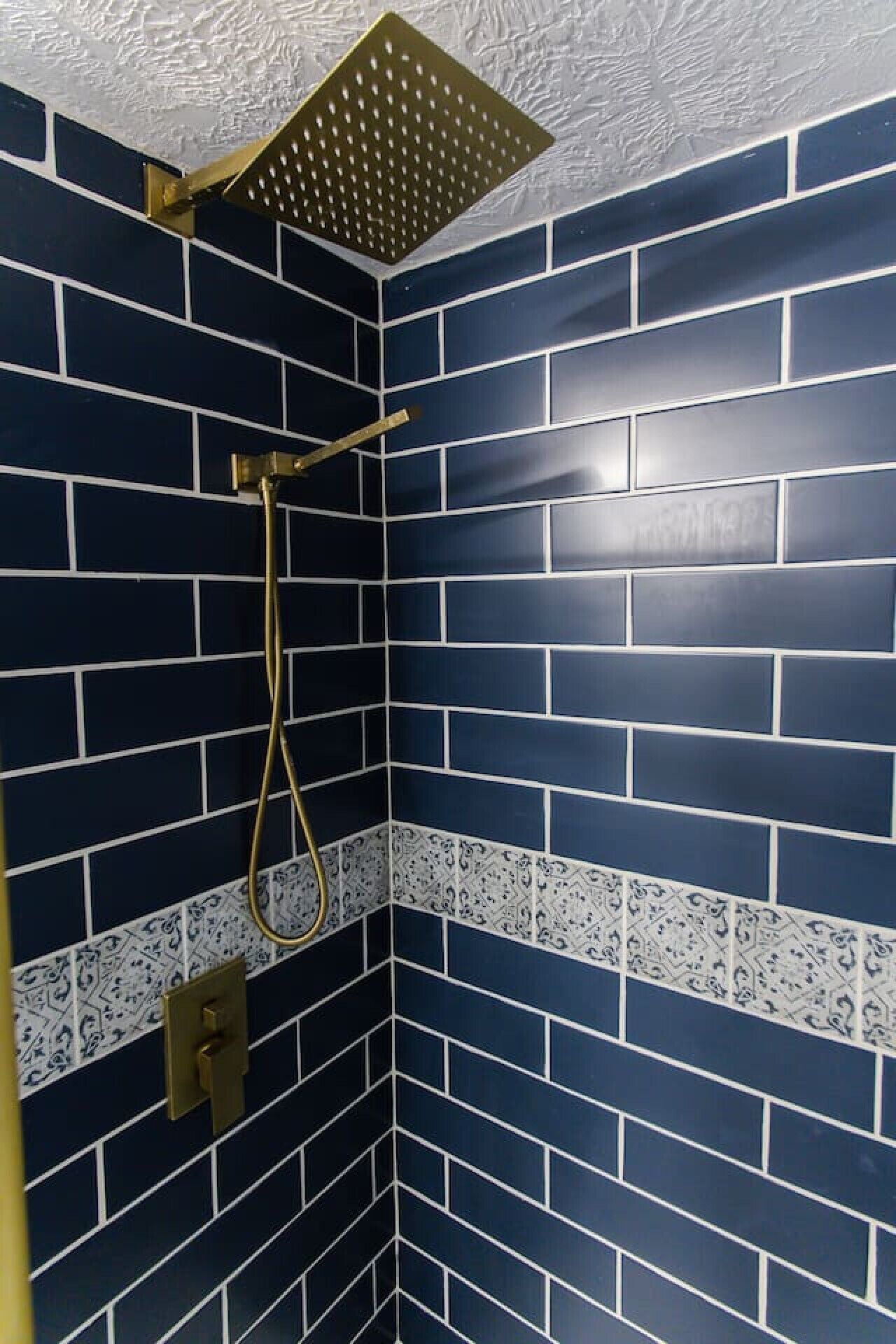 A shower with a gold showerhead and a gold handle, set in a corner with dark blue tiled walls and a white and blue patterned border.