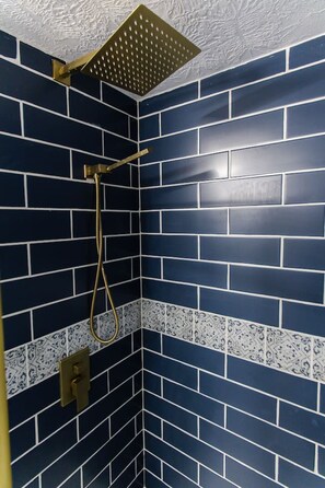 A shower with a gold showerhead and a gold handle, set in a corner with dark blue tiled walls and a white and blue patterned border. - Stylish, comfort-first 2BR/1BA (Sioux Falls)