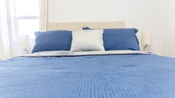 Een bed met een blauwe spreideken en twee kussens, één wit en één blauw, in een kamer met een wit hoofdeinde en een lamp ernaast.