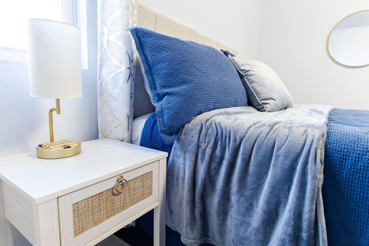 A neatly arranged bedroom with a bed, a bedside table with a lamp, and a wall-mounted mirror.