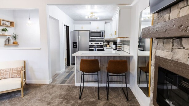 A modern kitchen with white cabinets, a central island with bar stools, a stone fireplace, and a dining area with a sofa and pendant lights. - Stylish, comfort-first 2BR/1BA (Sioux Falls)