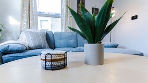A living room with a blue sofa, a round table, a potted plant, and a small side table with a patterned cushion. - Stylish, comfort-first 2BR/1BA (Sioux Falls)