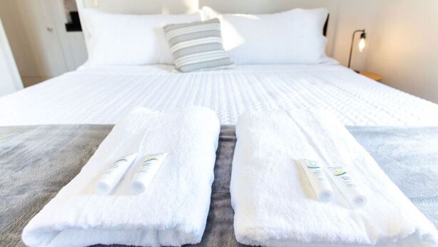 Ein ordentlich gemachtes Bett mit weißen Leinentüchern und zwei gefalteten Handtüchern mit Aufrollröhren auf einer grauen Fläche.