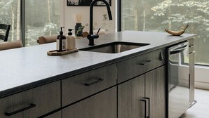 Una cocina moderna con una isla grande, un grifo negro y vistas a un bosque a través de amplios ventanales.