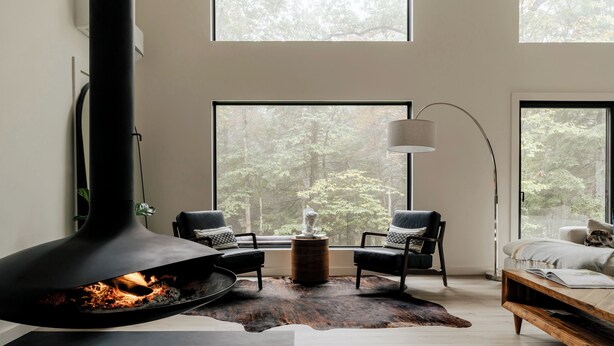 Una sala de estar moderna con una chimenea grande, dos sillones de cuero negro, una mesa de centro de madera y vistas a un bosque a través de amplios ventanales.
