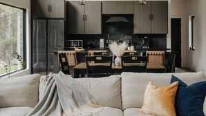 Una sala de estar moderna con un sofá, una mesa de centro y libros. Se observa un comedor con sillas y una mesa. La cocina es visible al fondo, con armarios y electrodomésticos.