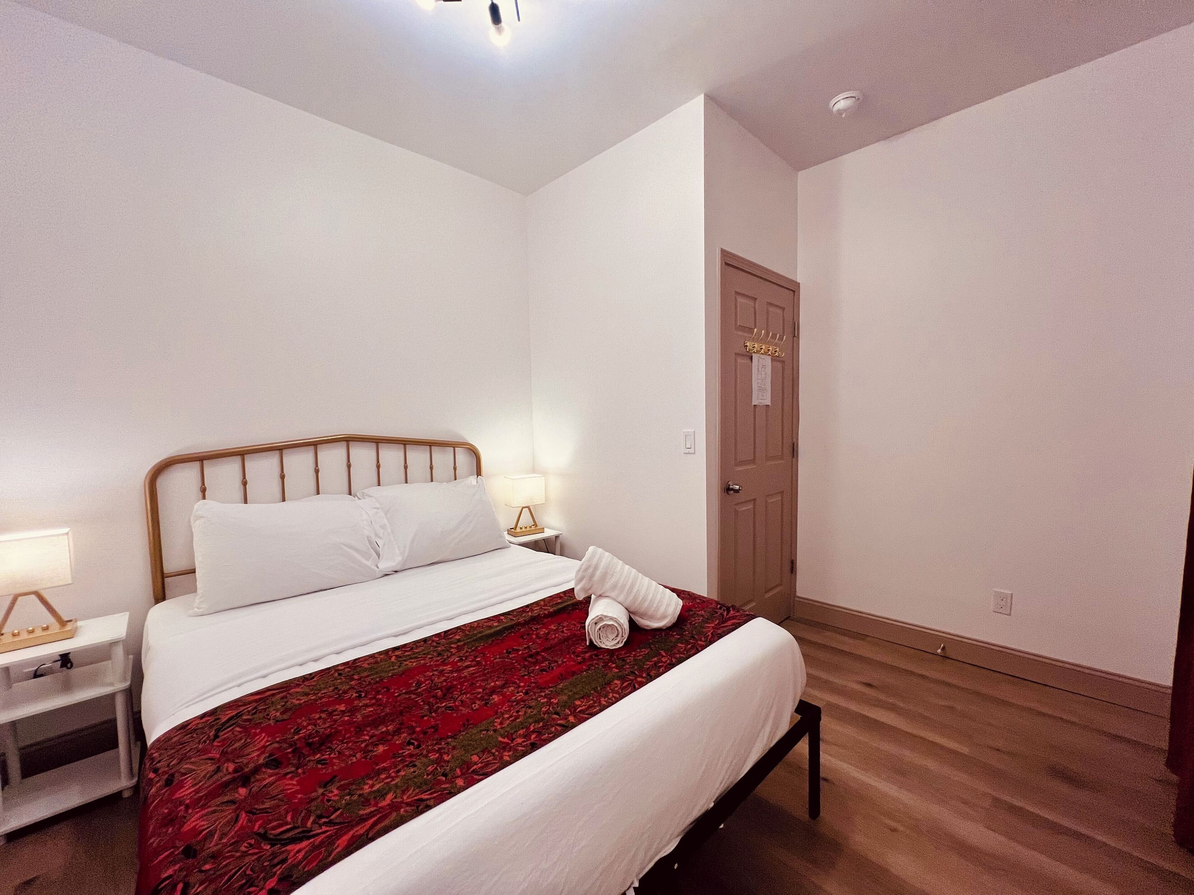 Een net opgemaakt bed met witte lakens en een rood geborreld sprei, een nachtkastje met een lamp, en een deur met een bordje.