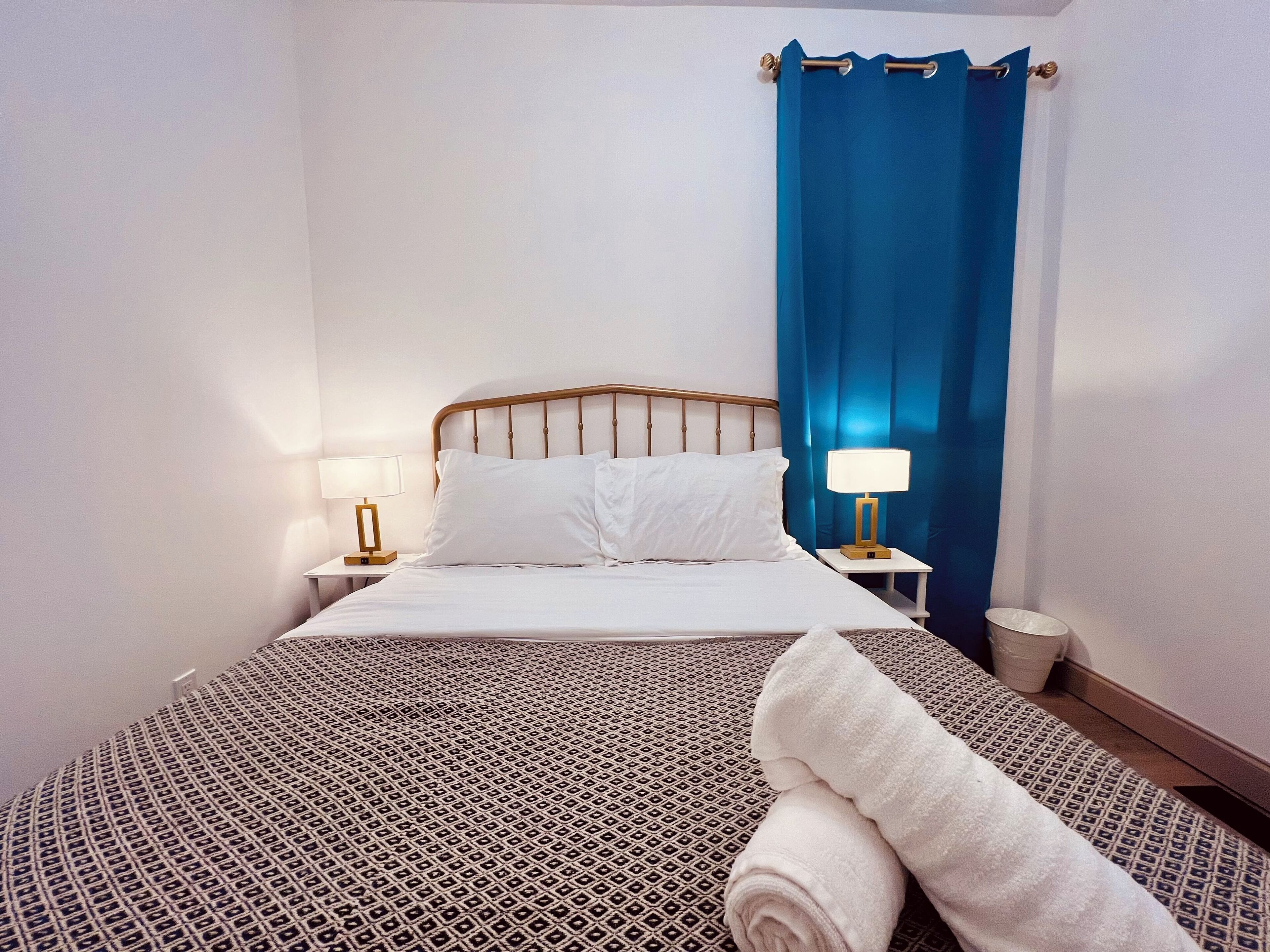 Una cama bien hecha con sábanas blancas, dos lámparas de noche y una cortina azul.