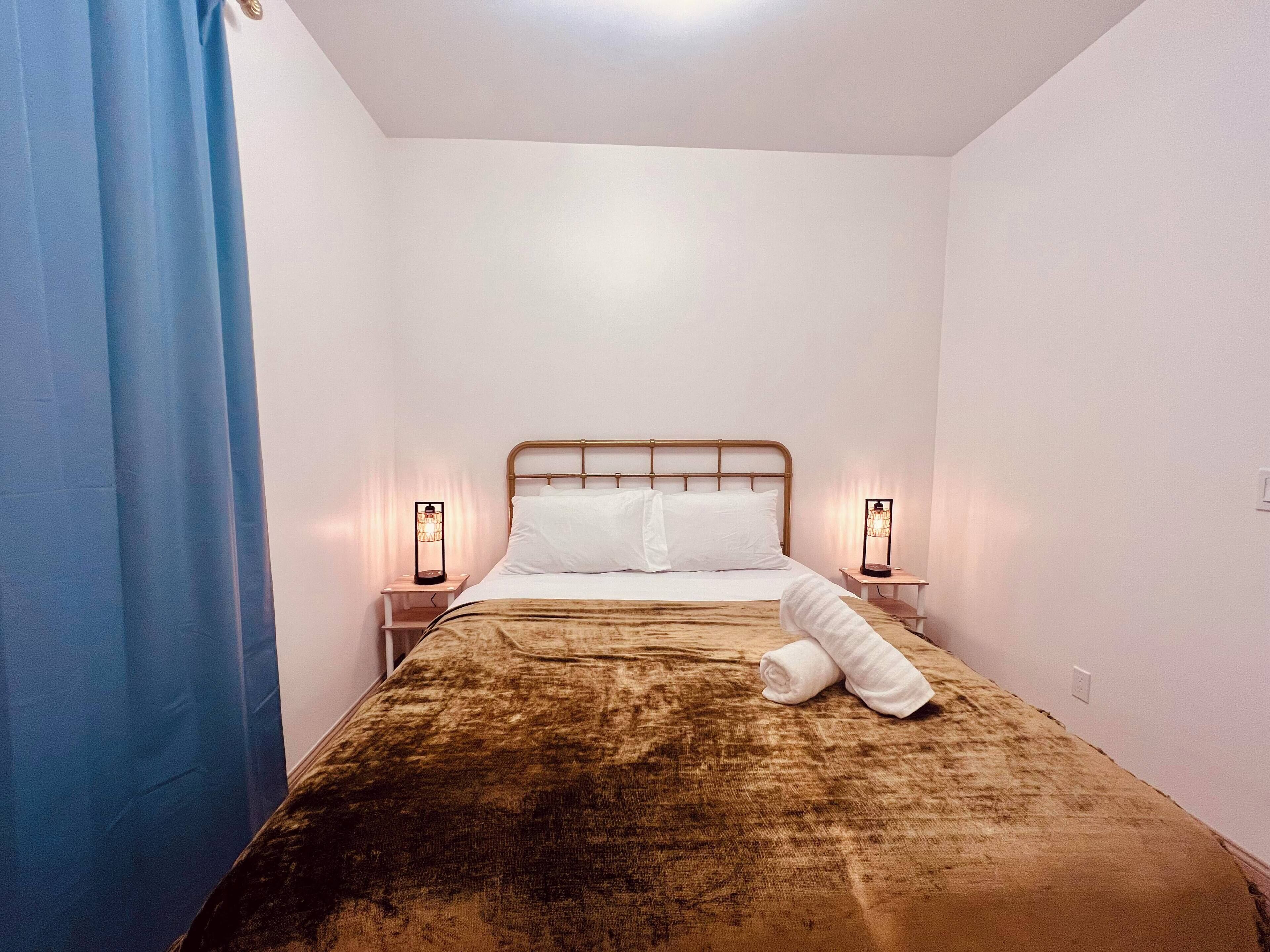Un lit simple avec une tête de lit, deux tables de chevet avec des lampes, un rideau bleu et une serviette pliée sur le lit.