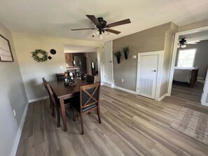 A dining area with a wooden table and chairs, a ceiling fan, and a kitchen in the background.
