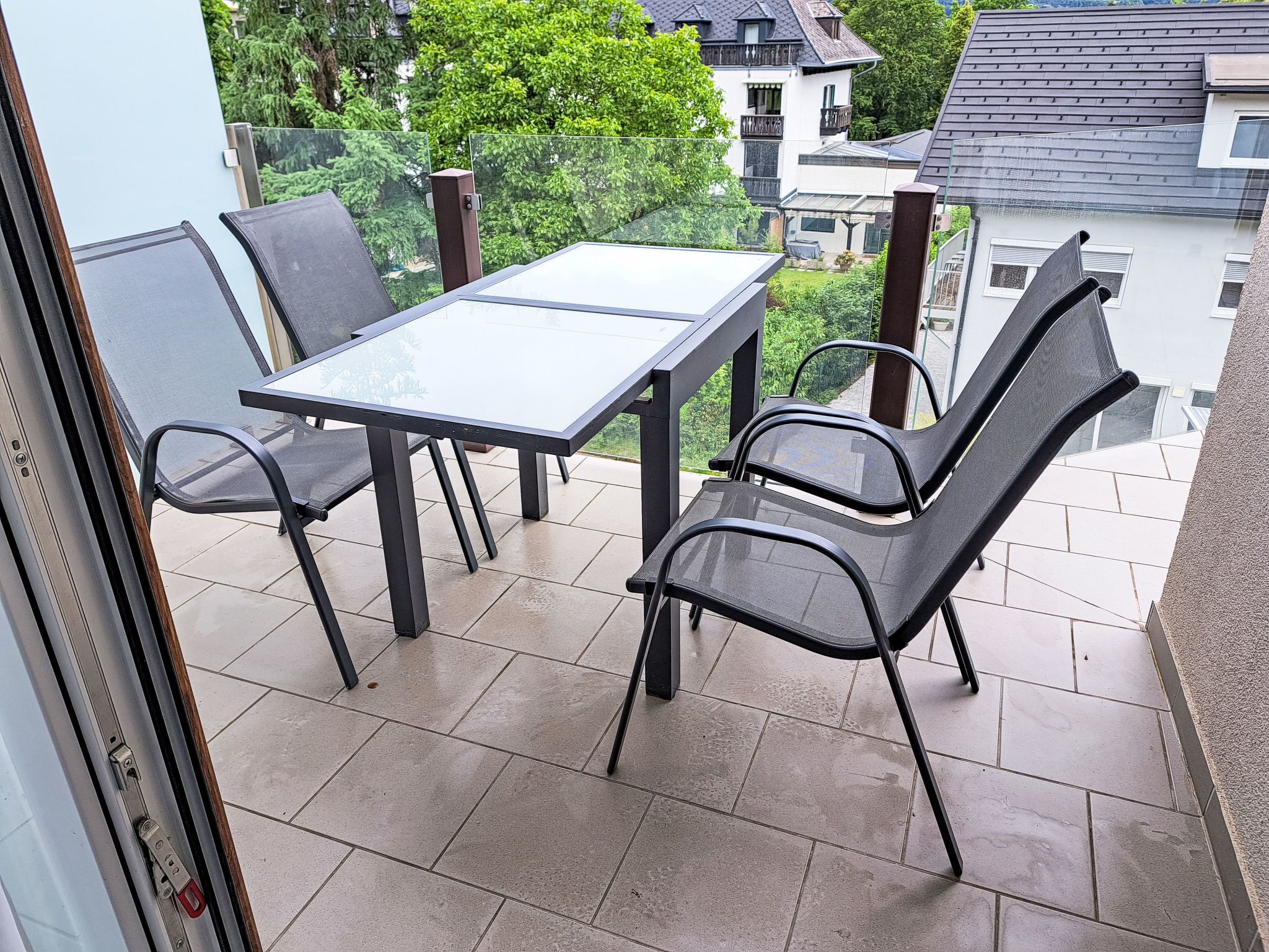 Un balcon doté d’une table en verre et de quatre chaises, donnant sur un quartier résidentiel avec des maisons et des arbres.