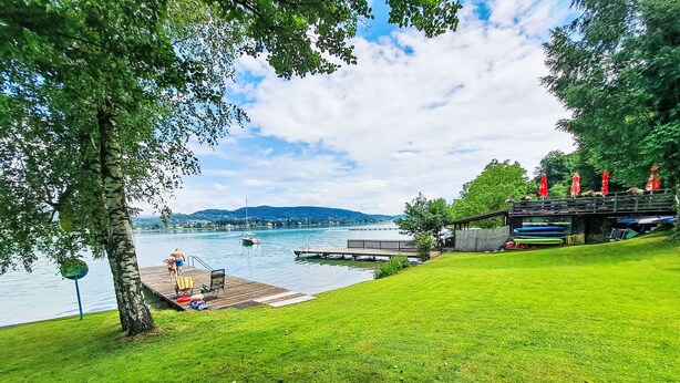 A lakeside view with a grassy area, a dock, and a building with red umbrellas. - Sunshine view 3 by Interhome (Reifnitz)