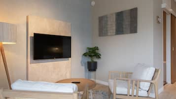 A modern living room with a flat-screen TV mounted on the wall, a potted plant, two white cushioned chairs, a round coffee table, and a wooden floor.
