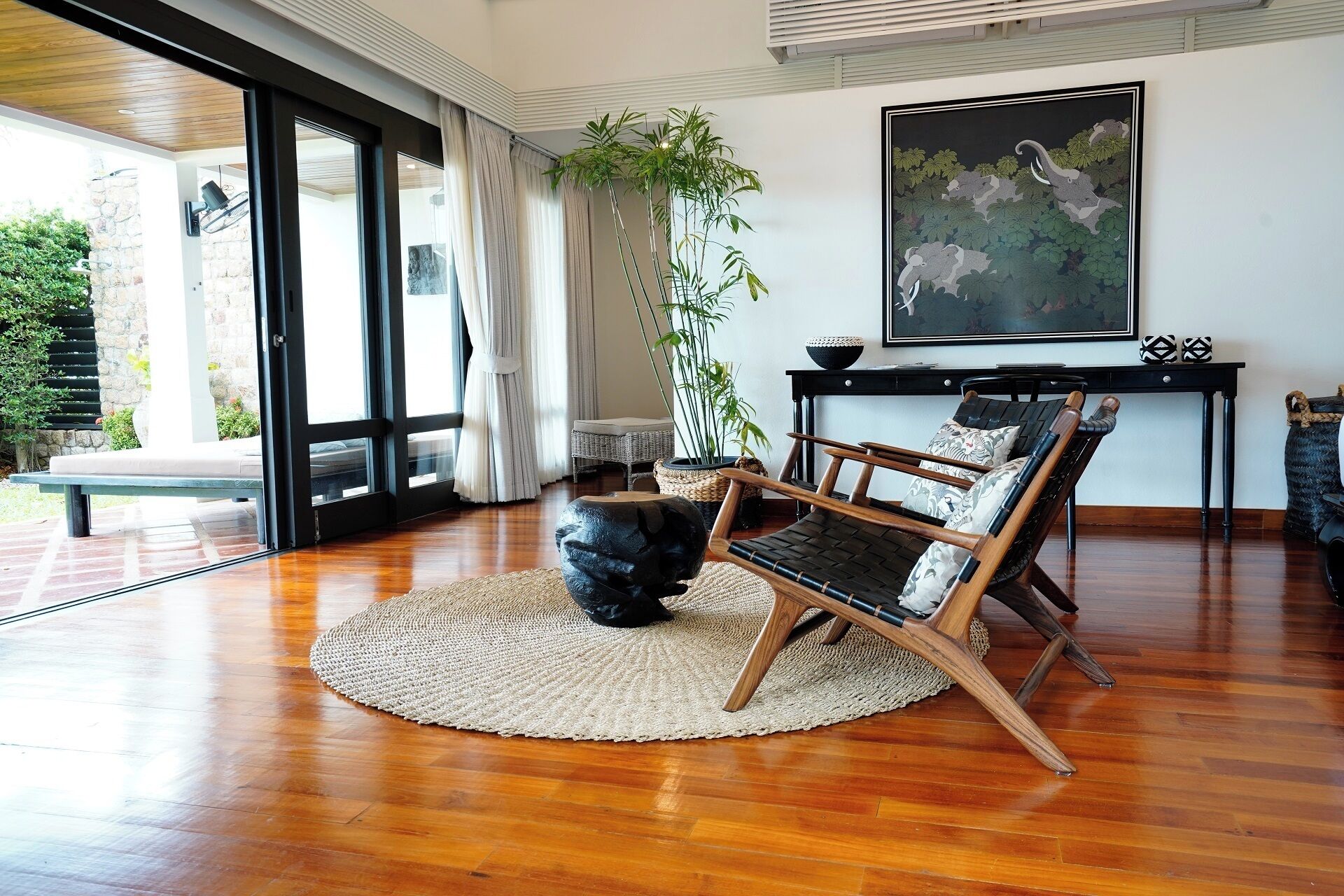 A spacious living room with a large window, a wooden floor, a black chair, a woven rug, and a painting on the wall.