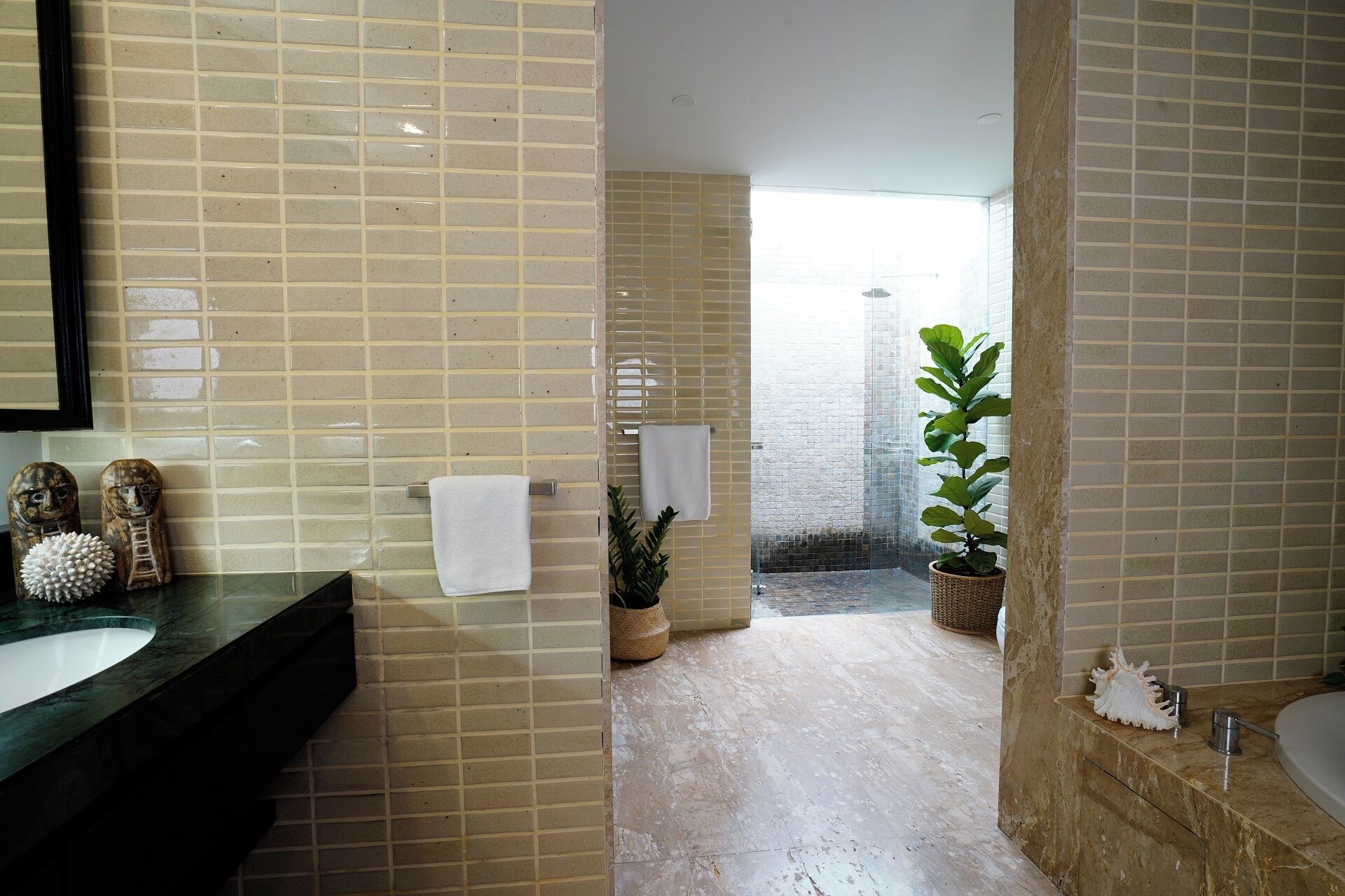 Une salle de bain avec des murs carrelés de couleur beige, un comptoir en marbre, un lavabo, un miroir, deux serviettes, une plante en pot et un espace douche.