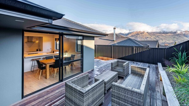 A modern house with a kitchen and dining area visible through glass doors, a wooden deck with wicker furniture, and a mountainous backdrop. - Mountain View Haven T (Wanaka)