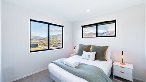 A bedroom with a large bed, two nightstands, a lamp, and two windows with mountain views. - Mountain View Haven T (Wanaka)