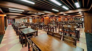 Un'ampia sala da pranzo con numerosi tavoli e sedie in legno, soffitti alti con travi a vista e illuminazione al neon.