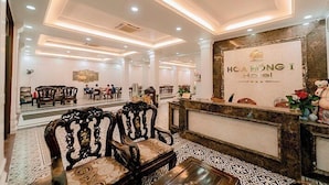 Hall d'hotel con poltrone in legno intagliate, una reception e un grande specchio che riflette l'ambiente.