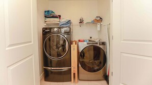 Et lille vaskerum med to stablede vaskemaskiner og tørretumblere, en hylde med foldet tøj, og en kost lænet op ad væggen.