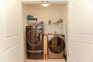 A compact laundry room with two stacked washers and dryers, a small shelf with folded clothes, and a broom leaning against the wall. - Sojourns Beach Views & Pool Family Home Sleeps 9 (Virginia Beach)