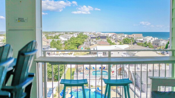 Udsigt fra en balkon med pool og stole, der kigger ud over et boligområde med huse og en klar himmel.