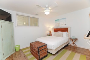 Quarto com cama, ventilador de teto, janela com persianas, lixeira verde e um baú de madeira.