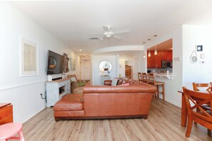 Sala de estar com sofá de couro marrom, mesa de jantar de madeira e cadeiras, cozinha com armários vermelhos e um ventilador de teto.