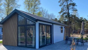 A modern single-story house with large glass windows, a sloped roof, and a patio area with outdoor furniture. - Nice Chalet in a Holiday Park, Adjacent to the Hoge Veluwe National Park (Arnhem)