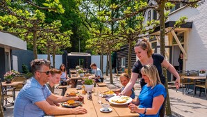 An outdoor dining area with wooden tables and benches, surrounded by trees and a building with a white facade. - Nice Chalet in a Holiday Park, Adjacent to the Hoge Veluwe National Park (Arnhem)