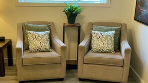 A living room with two armchairs, a small table, a potted plant, and a large window with a view of a house and greenery. - Cosy Hideaway in the Heart of Tofino (Tofino)