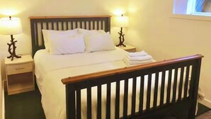 A neatly made bed with white linens, two bedside tables with lamps, and a window with blinds. - Cosy Hideaway in the Heart of Tofino (Tofino)