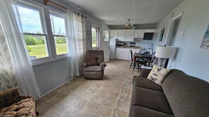 Una sala de estar con un sofá marrón, un sillón estampado y un comedor con mesa y sillas. Se ve una cocina con armarios y electrodomésticos al fondo.