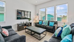 A spacious living room with a gray sofa set, a coffee table, and a TV mounted on the wall. There are windows with blinds, a ceiling fan, and a small side table with a lamp.