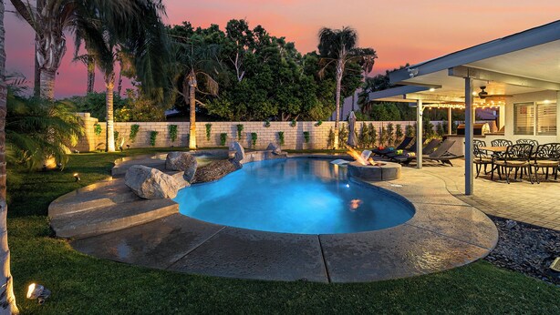 Piscina con borde de piedra, rodeada de césped y palmeras, con un porche cubierto y mobiliario de exterior.