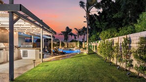 Un patio trasero con piscina, cocina al aire libre y un porche con luces colgantes.
