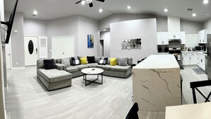 Una sala de estar moderna con un sofá modular, una mesa de centro redonda y una cocina con armarios y electrodomésticos blancos.