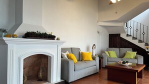 Een woonkamer met een openhaard, twee grijze banken met gele en groene kussens, een houten salontafel en een trap met een leuning.