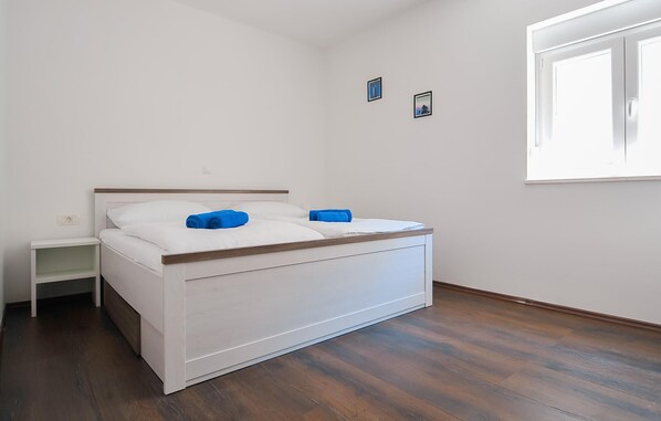 Une chambre avec un lit simple, une tête de lit en bois, du linge de lit blanc, une petite table de chevet et une fenêtre avec des stores.