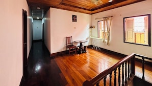 A room with wooden flooring, a ceiling fan, a table with an ironing board, a chair, and a window with shutters. - Silikhane Guest House (Luang Prabang)