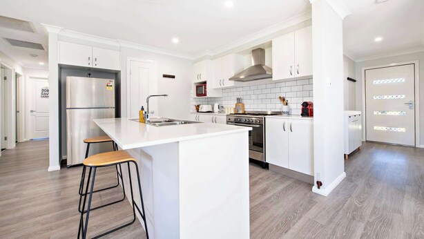 A modern kitchen with white cabinetry, a stainless steel refrigerator, a built-in oven, and a large island with stools. - Magnolia Entertainer. Firepit & Pets. Dubbo Family (Dubbo)
