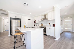 A modern kitchen with white cabinetry, a stainless steel refrigerator, a built-in oven, and a large island with stools. - Magnolia Entertainer - Firepit, Pets Welcome (Dubbo)