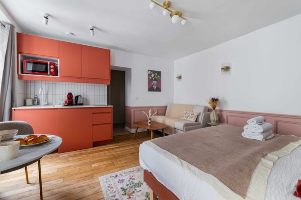 A modern hotel room with a kitchenette, a bed with a beige blanket, a sofa, and a small table with a vase of flowers.