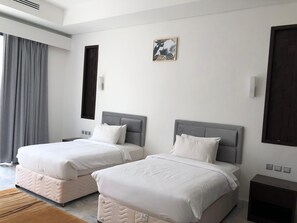 A hotel room with two beds, a large window with curtains, a wall-mounted picture, and a small bedside table. - The Palace Hotel Salalah (Salalah)